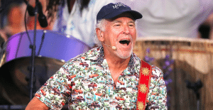 This photo shows Jimmy Buffett performing onstage with his guitar, and mouth open in excitement.